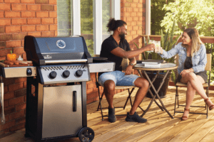 Man and woman sit on their patio deck in the background giving a cheers to each other with a BBQ grill in the foreground